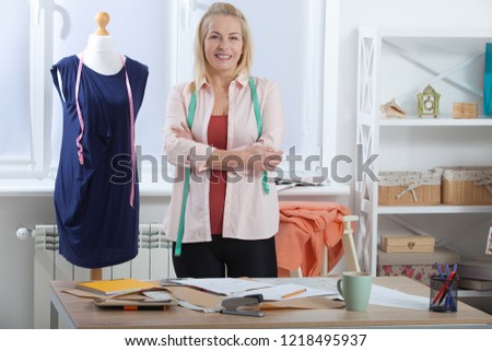 Successful woman designer clothes, tailor in studio in full growth. Beautiful woman looking at the camera, standing near table in the workshop with clothes. 