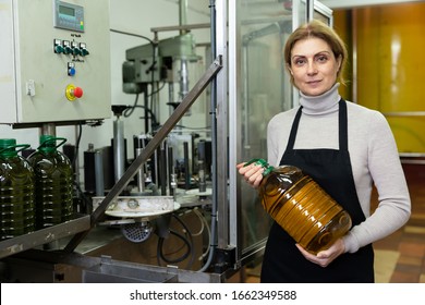 Successful woman owner of small olive oil factory standing near bottling line with plastic bottle filled of finished product in hands