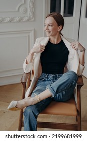 Successful Woman Is Sitting In Vintage Armchair With Her Legs Crossed. Portrait Of Stylish Young Adult European Lady In White Jacket Posing In Studio. Concept Of Elegance And Confidence.