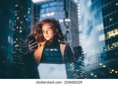 Successful woman using smartphone outdoors while standing near skyscraper at night. 