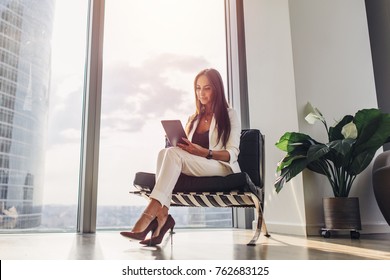 Successful Woman Wearing Suit Sitting On Armchair Using Tablet Computer At Her Loft Apartment In City Center