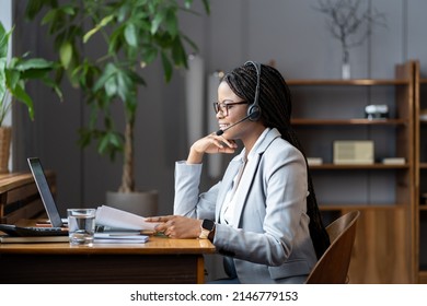 Successful Young African American Businesswoman Sitting At Table In Cozy Home Office Managing Remote Team Of Workers, Black Female Freelancer Using Wireless Headset While Working Remotely On Laptop