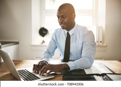 Successful young African executive sitting at his desk in an office smiling and working on a laptop