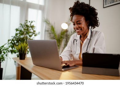 Successful Young Afro American Female Doctor In White Medical Uniform And Stethoscope Busy Working On Laptop In Modern Hospital, Smiling Woman Nurse Or GP Typing On Computer Consult Patient Online.