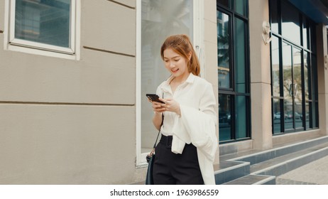 Successful Young Asia Businesswoman In Fashion Office Clothes Using Smart Phone And Typing Text Message While Walking Alone Outdoors In Urban Modern City In The Morning. Business On The Go Concept.