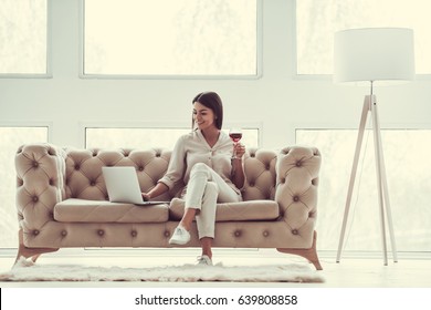 Successful Young Beautiful Woman Sitting On A Sofa In The Living Room. Using Laptop And Drinking Red Wine.