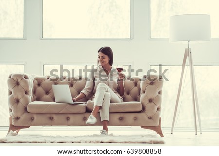 Successful young beautiful woman sitting on a sofa in the living room. using laptop and drinking red wine.