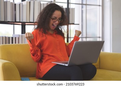 Successful Young Black Woman Wearing Orange Shirt Exciting And Using Laptop For Video Call On Sofa At Home. Business Woman And Freelance Work From Home In Spreading Of Coronavirus Covid-19.