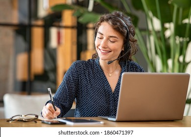 Successful young business woman working on laptop with headphones in a call center as a consultant. Happy businesswoman with headset translating and writing notes by listening to audio course.