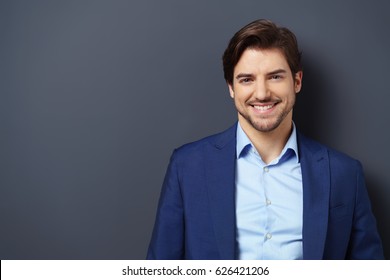 Successful Young Businessman In A Blue Suit With A Lovely Genuine Smile Posing Against A Dark Grey Background With Copy Space