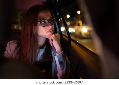 Successful young businesswoman in fashion office clothes working late and using smartphone and looking trough the window while sitting in a backseat of car in urban modern city during night. - Shutterstock ID 2233476897