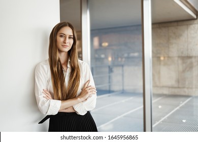 Successful young businesswoman lean wall and cross hands on chest, smiling confident and assertive, express readiness, just signed big financial deal for her company, standing inside office