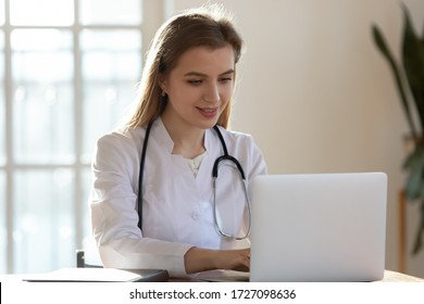 Successful Young Caucasian Female Doctor In White Medical Uniform And Stethoscope Busy Working On Laptop In Modern Hospital, Smiling Woman Nurse Or GP Typing On Computer Consult Patient Online