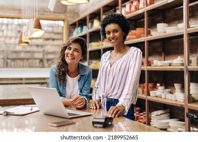 Successful young ceramists smiling at the camera while working in their store. Happy female entrepreneurs using a laptop together. Two young businesswomen running a creative small business.