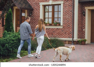 Successful Young Couple Walking With A Dog Near Home. Beautiful Family Moving To Their New House.