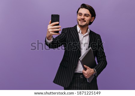 Successful young gentleman in white shirt, black striped suit and trendy glasses making selfie with folders and smiling. Business concept isolated over violet background