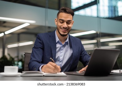 Successful young middle eastern businessman working at modern office, sitting at workdesk in front of laptop, typing on keyboard and taking notes, planning his working day, copy space
