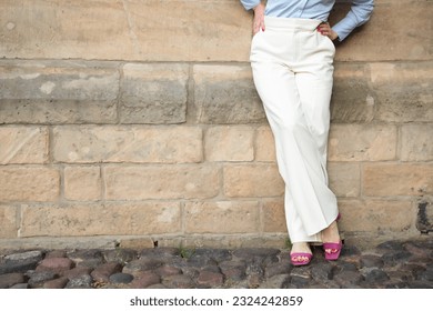 a successful young woman in white trousers and a blue shirt. the woman is leaning against the wall. A shortened shot of a woman's legs in classic trousers. copy space - Shutterstock ID 2324242859