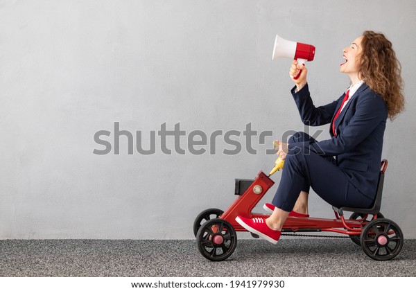 Successfull businesswoman driving\
toy car outdoor. Funny young woman shouting through megaphone\
against concrete wall background. Business srart up and winner\
concept