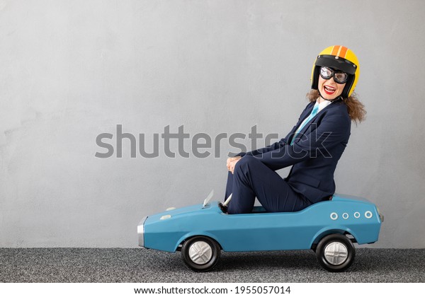Successfull businesswoman driving toy car\
outdoor. Funny young woman against concrete wall background.\
Business srart up and winner\
concept