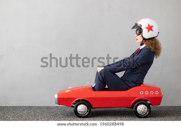 Successfull businesswoman driving toy car\
outdoor. Funny young woman against concrete wall background.\
Business srart up and winner\
concept