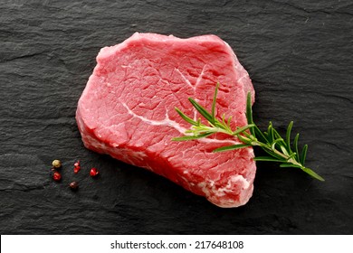 Succulent Cut Of Raw Lean Rump Steak Waiting To Be Cooked With A Sprig Of Fresh Rosemary For Seasoning And Whole Peppercorns, View From Above On A Dark Kitchen Counter