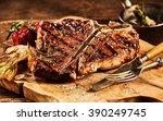 Succulent grilled large t-bone steak garnished with herbs, tomato and salt with fork and knife beside it on cutting board