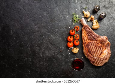 Succulent grilled tomahawk beef steak on the bone with red wine, seasonings, fresh rosemary and grilled vegetables on a black background, top view