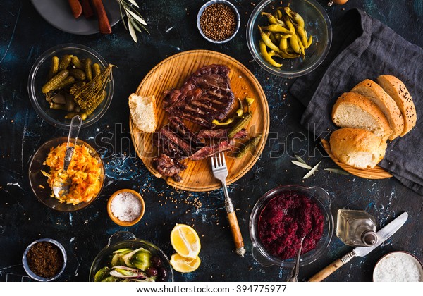 Succulent thick juicy portions of grilled fillet\
steak served with variety veggies dips, fermented veggies, cucumber\
and pepper marinated, bread buns on a old vintage table  Authentic\
dinner party.