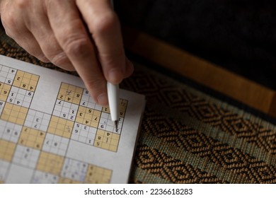 Sudoku brain game with numbers on a newspaper, holding a pen in a cosy warm home. Great exercise for elderly people to keep a healthy active mind, reduce alzheimer and dementia illness