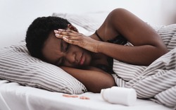 Suffering From Pain And Migraine. Ailing Young African American Lady With Closed Eyes Presses Hands To Temples And Suffers From Headache Lies On Bed In Interior Of Bedroom With Jar Of Pills, Close Up