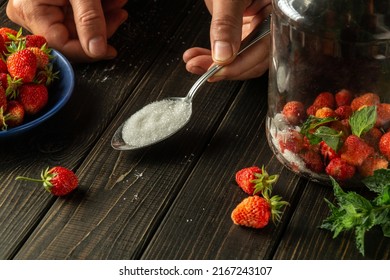 Sugar is added to a jar of ripe strawberry and mint compote. Hands of chef with spoon on kitchen table.