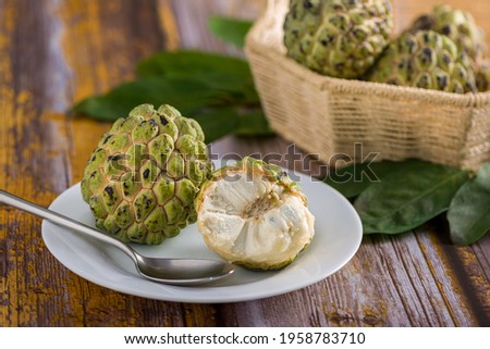 Sugar apple on a white plate with stainless steel spoon, ready to be eaten. In addition to other sugar apples in a basket on a blurred background on an aged yellow wooden table.