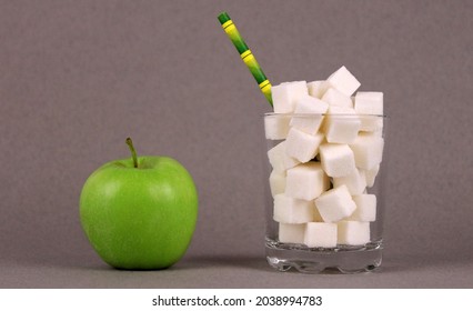 Sugar Cubes In A Glass With A Straw And A Green Apple On A Gray Background. Healthy Food Versus Unhealthy Sugary High-calorie Drinks