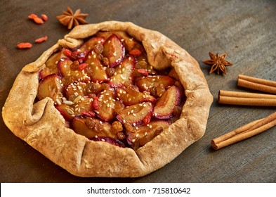 Sugar free pie with plums, raisins, goji berries and sesame on brown wooden background. Pie with plums decorated with dried goji berries, anise stars and sticks of cinnamon close up. Top view