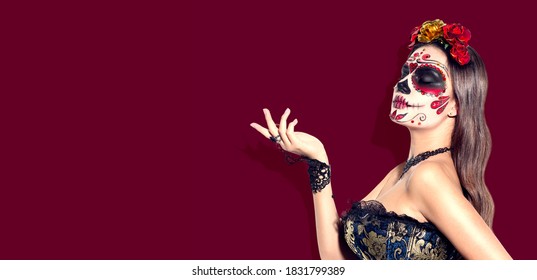 Sugar skull makeup. Halloween party, traditional Mexican carnival, Santa Muerte. Beautiful young woman costume, painted face. Model girl pointing hand, on red background. Calavera Catrina. 