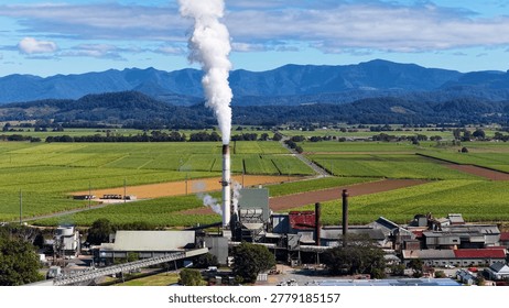 A sugarcane processing plant emits white smoke amidst lush green fields and distant mountains.