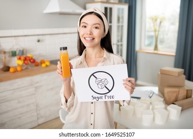 Sugar-free orange juice. Beautiful dark-haired young girl standing in the kitchen holding an orange juice and a poster without sugar, inspired full of energy.