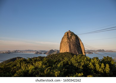 Sugarloaf Mountain In Guanabara Bay In Urca Neighbourhood At Sunset With Warm Sky Overhead, Mountains In The Background & Green Forest Trees In The Foreground In Rio De Janeiro, Brazil, South America