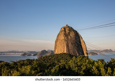 Sugarloaf Mountain In Guanabara Bay In Urca Neighbourhood At Sunset With Warm Sky Overhead, Mountains In The Background & Green Forest Trees In The Foreground In Rio De Janeiro, Brazil, South America