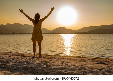 Suggestive shot of rear view of thoughtful woman silhouette in spiritual pose with open arms looking at sunset or sunrise barefoot on sea sand or ocean beach. Find yourself in contact with nature