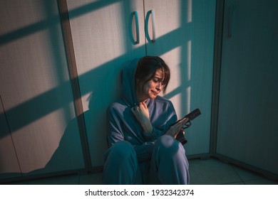 Suicidal woman sitting on the floor in the dark, holding a gun and crying, thinking about committing suicide; scared woman victim of domestic violence holding a gun for self-defense