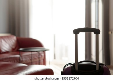 A Suitcase Standing By The Window In The Living Room Near A Large Leather Red Sofa And An Empty Table. The Concept Of Moving And Starting A New Life In A New Apartment. No People Interior