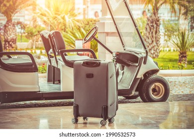 A Suitcase Standing Near Electrical Hotel Passenger Shuttle Bus In Front Of Blurry Palm Trees. Summer Holiday Concept With Golf Car Service And Luggage.