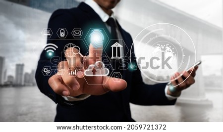 A suit-clad Asian man sits at a desk, his left hand holding a cellphone. His right hand is pointing to a hologram that depicts business icons in a hitech circle. Blurred black background