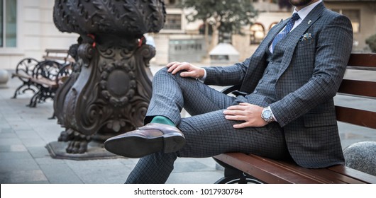 Suited man sitting on bench outdoors with his leg crossed and with his hands on his legs
