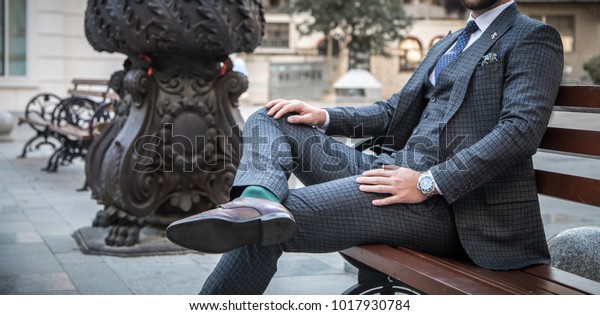 Suited man sitting on bench outdoors\
with his leg crossed and with his hands on his\
legs