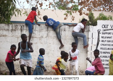 Sukuta, The Gambia, Africa, May 18, 2017:   Horizontal Photo Of A Group African Children In Colorful  Clothes, Climbing A White Wall With Written School  Mission Statement,  Outdoors On A Sunny Day 