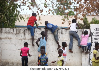 Sukuta, The Gambia, Africa, May 18, 2017:   Horizontal Photo Of A Group African Children In Colorful  Clothes, Climbing A White Wall With Written School  Mission Statement,  Outdoors On A Sunny Day 