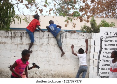 Sukuta, The Gambia, Africa, May 18, 2017:   Horizontal Photo Of A Group African Children In Colorful  Clothes, Climbing A White Wall With Written School  Mission Statement,  Outdoors On A Sunny Day 